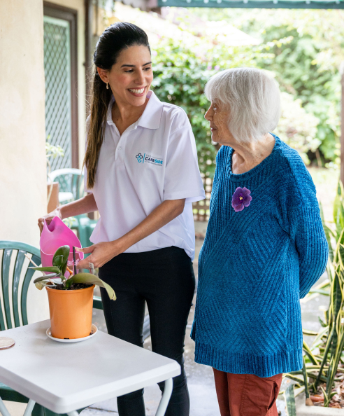 A caregiver from The CareSide waters a plant while standing next to her delighted home care client.