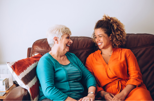 Home care client and her caregiver share a laugh while sitting on a couch.