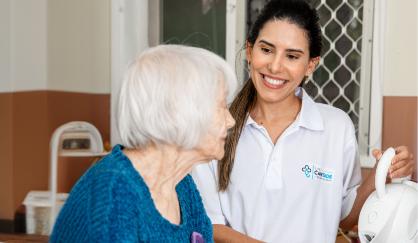 A caregiver from The CareSide smiles while standing at the kitchen counter with her client and pouring cups of tea.