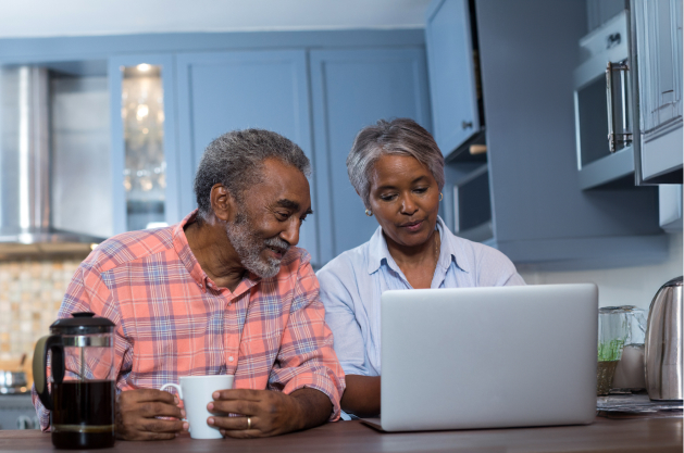 An older couple looks at a computer while sitting together in the kitchen.