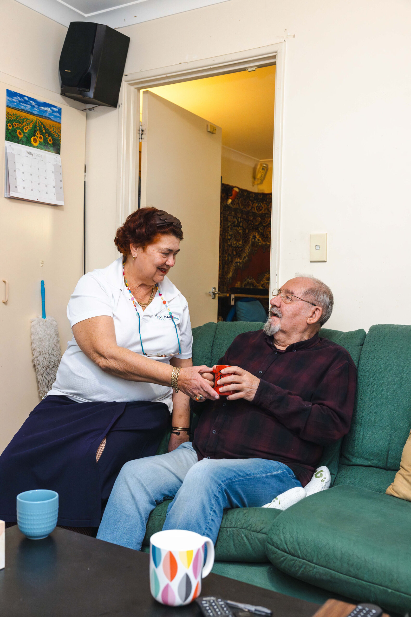 Home care in Darwin recipient sits on a sofa and accepts a cup of tea from his friendly caregiver from The CareSide.