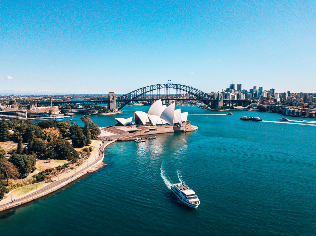 An aerial photo of Sydney Harbour shows a boat moving across the water with Sydney Opera House and Sydney Harbour Bridge in the background. Explore The CareSide's aged care jobs in Sydney.