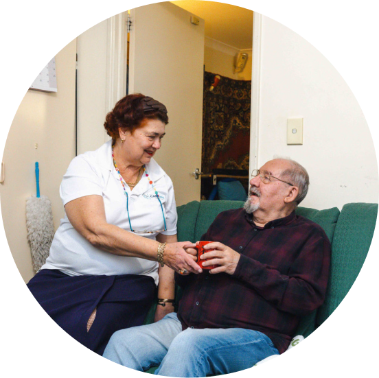 A caregiver from The CareSide handing a cup to her client sitting on the sofa.