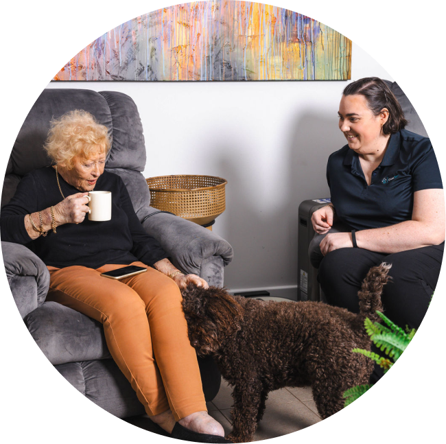 While receiving aged care services in Melbourne, a woman sips a cup of tea and pets her dog as her caregiver smiles nearby.