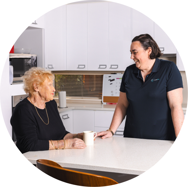 A smiling caregiver from The CareSide provides home care services in Sydney while chatting with her client across the kitchen counter.