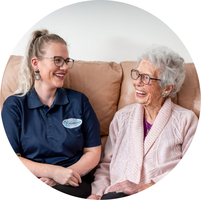 A caregiver from The CareSide provides home care services in Central Coast while sitting on a sofa and laughing with her happy client.