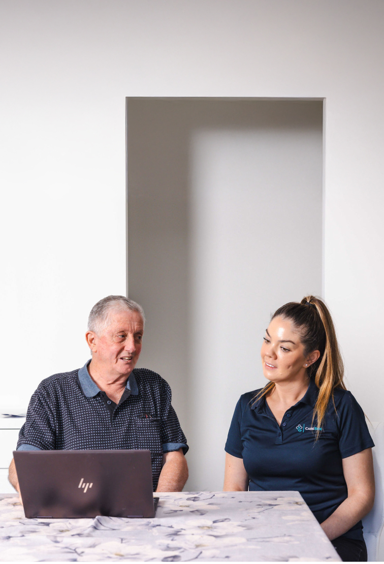 A caregiver from The CareSide provides home care services while sitting at a table with her client and helping him use a computer.