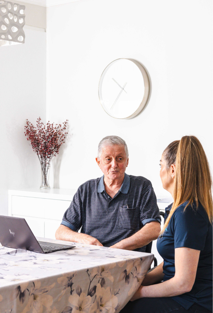 While sitting at a table, a Sydney aged care recipient has a conversation with his caregiver from The CareSide.