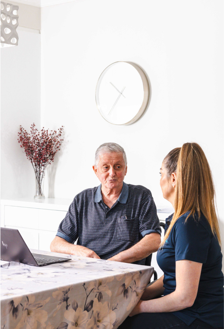 While sitting at a table, an aged care recipient has a conversation with his caregiver from The CareSide.