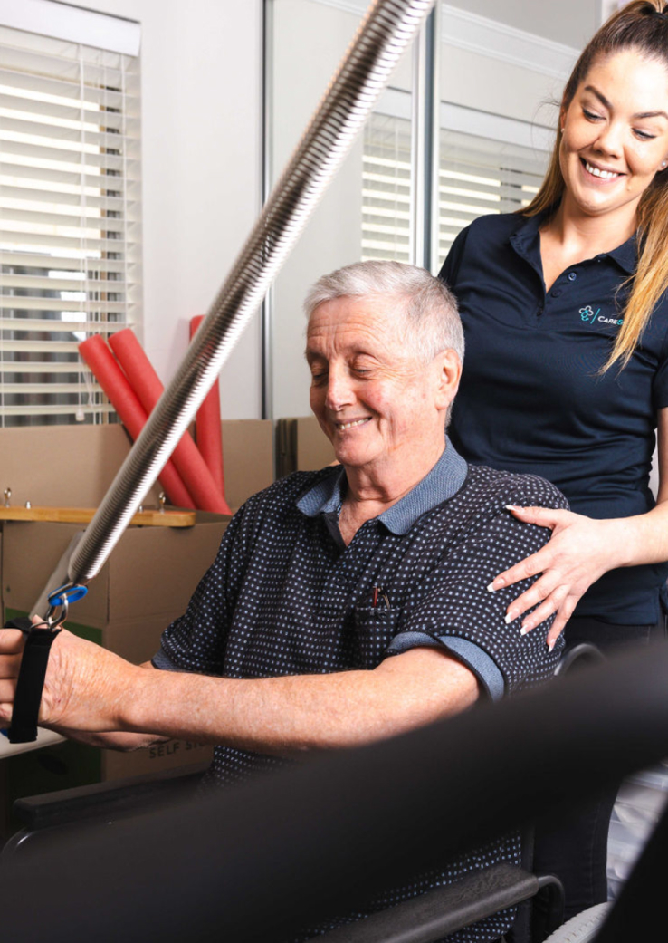 Home nursing care client smiles while performing rehabilitation exercises with assistance from a nursing care professional.