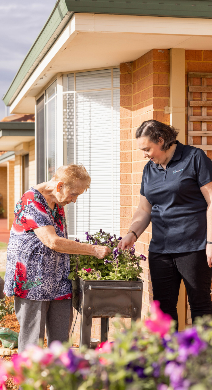 Companion care client works on a flower bed outside with assistance from her caregiver.