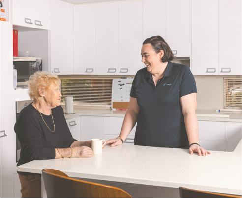 Aged care recipient stands at the kitchen counter while talking with her smiling caregiver.