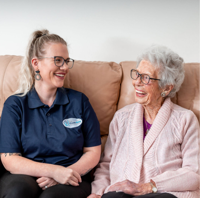 Home care client and her caregiver from The CareSide smiling at each other while sitting on a sofa.