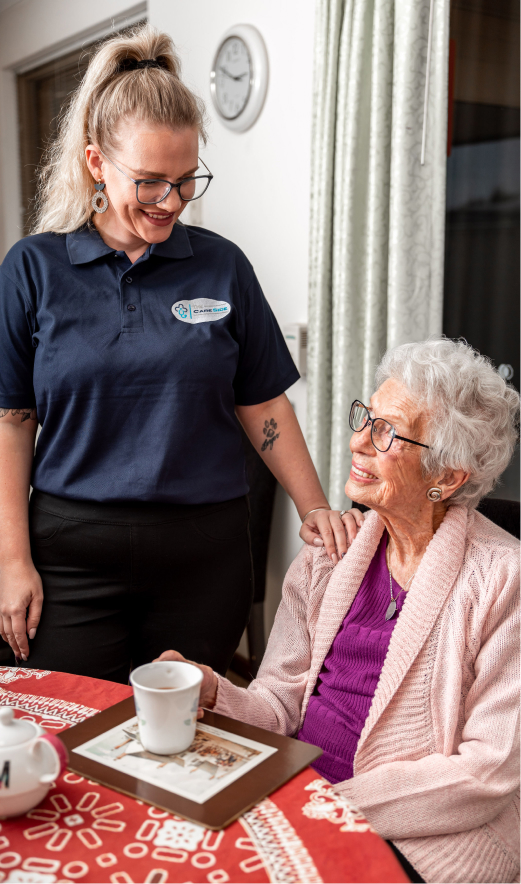 Respite care recipient sitting at a table and smiling at her caregiver standing next to her.