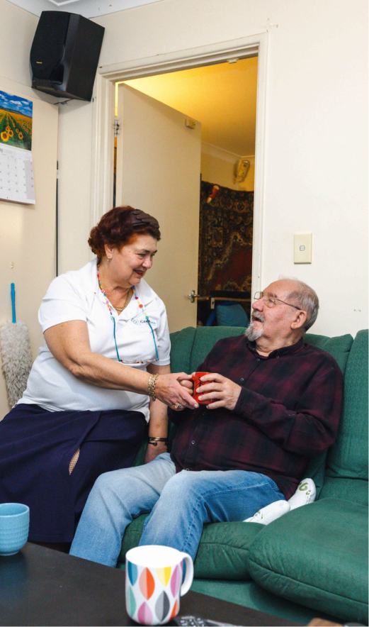 Home care client sitting on a sofa and accepting a cup of tea from his friendly caregiver.