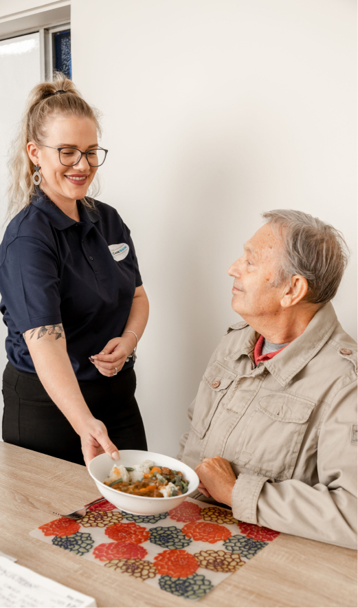 Post-hospital care patient sits at a table as his caregiver from The CareSide sets down a plate of food.