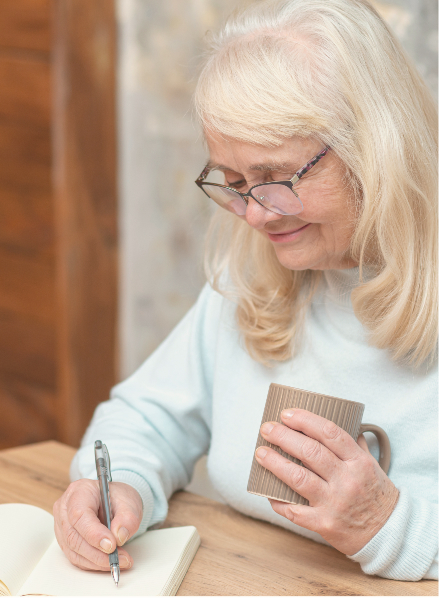 Woman sitting at a table holding a mug and writing in a notepad.